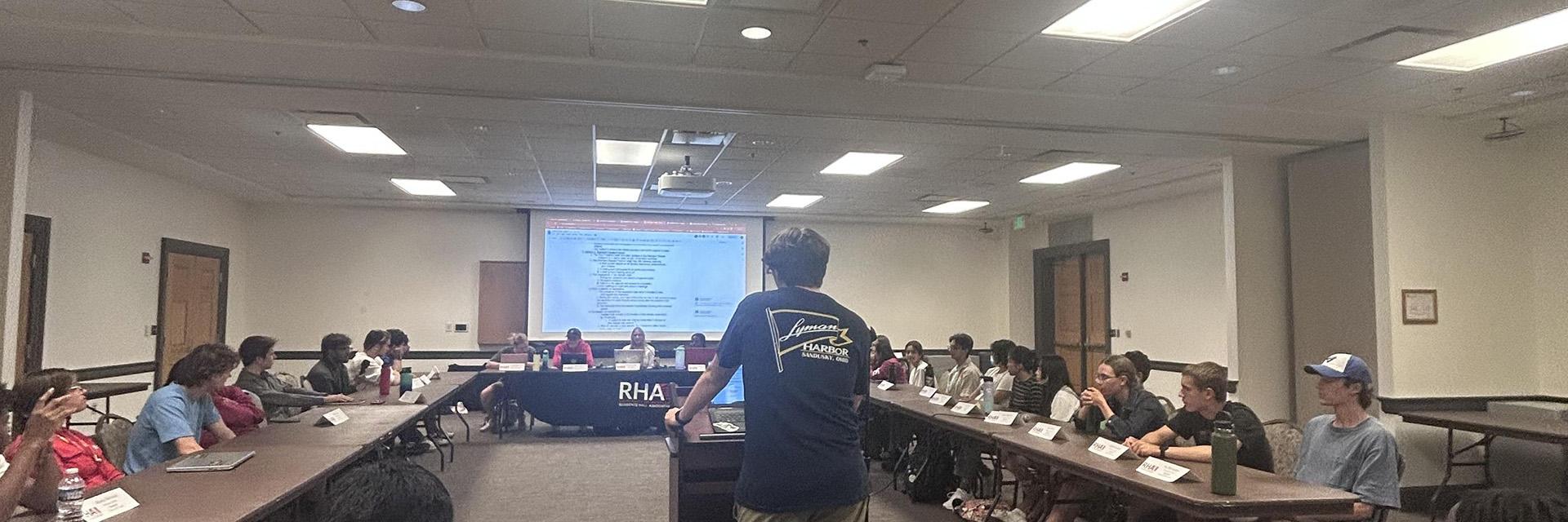 Students sitting at long tables in a U-shape for an RHA committee meeting with a projector screen.