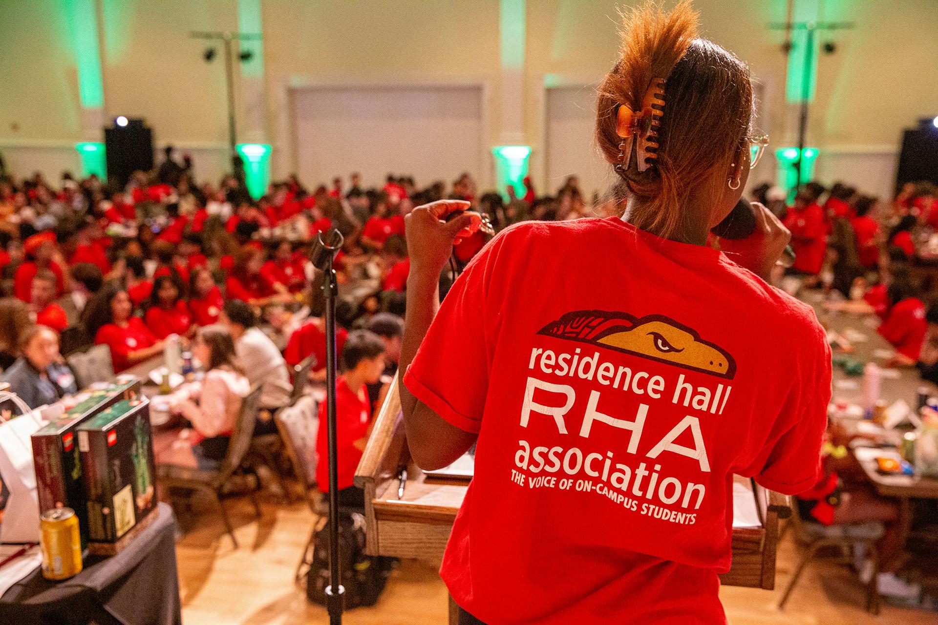 A speaker in a red RHA shirt faces a large crowd of students during a bingo event in a ballroom.