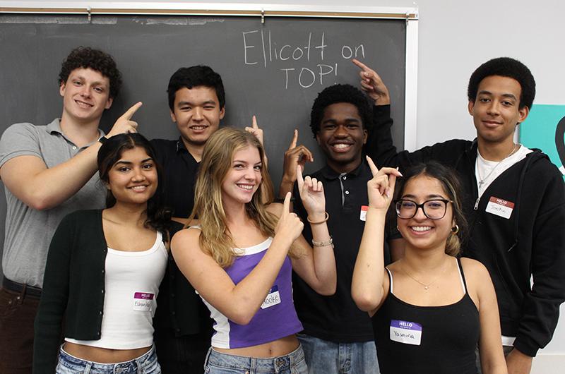 Seven RHA students smile and point toward a chalkboard behind them.