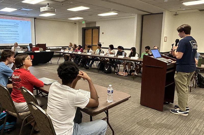 A student speaks at a podium to a seated group of students in a large conference room during an RHA meeting.