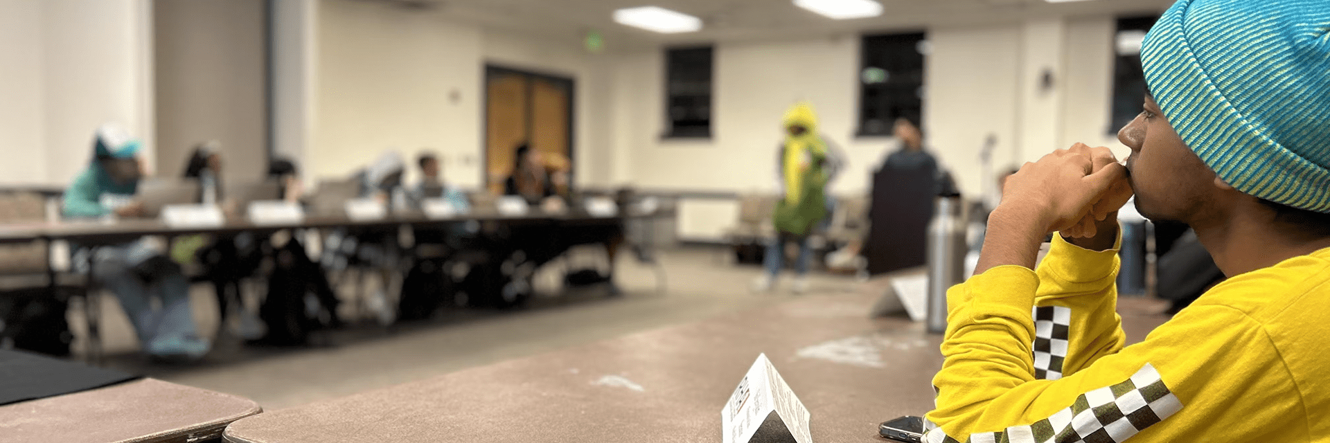A profile view of a student in a yellow shirt watching an RHA meeting in a large conference room.