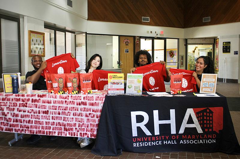 Four students hold up red "Leonardtown" shirts at an RHA information table in the Leonardtown Community Center.