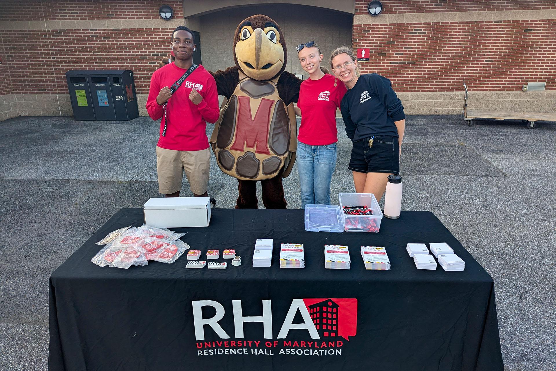 Three students and the Testudo mascot pose behind an RHA information table at the Big Show event during Fall Welcome.