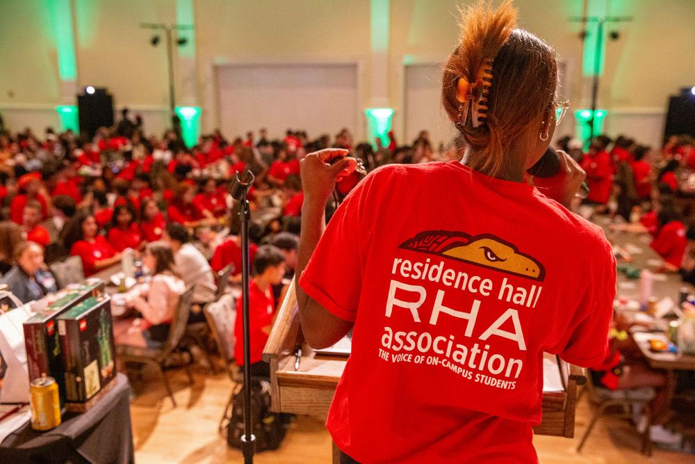 A speaker in a red RHA shirt faces a large crowd of students during a bingo event in a ballroom.